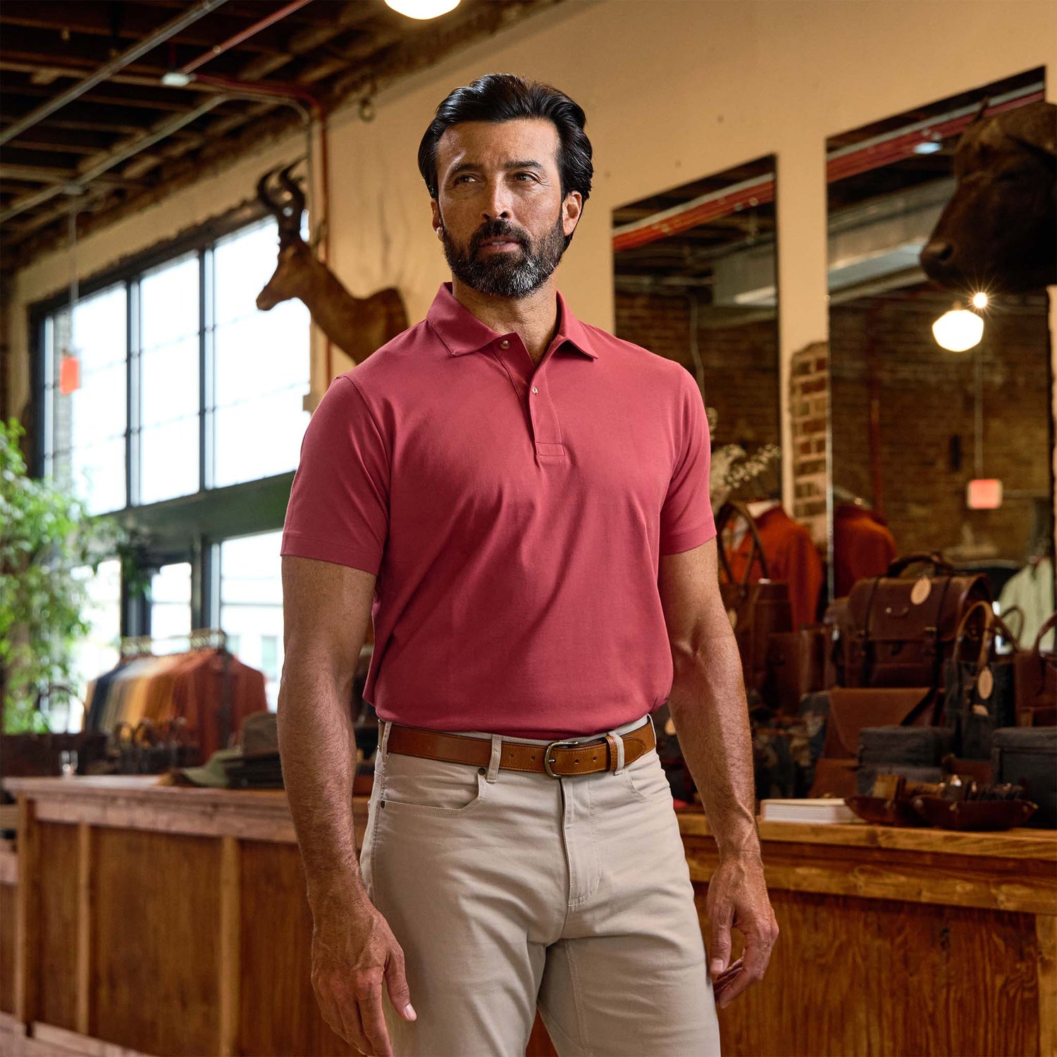 Man wearing a red polo shirt and beige pants stands inside a rustic store with leather goods and taxidermy on display.