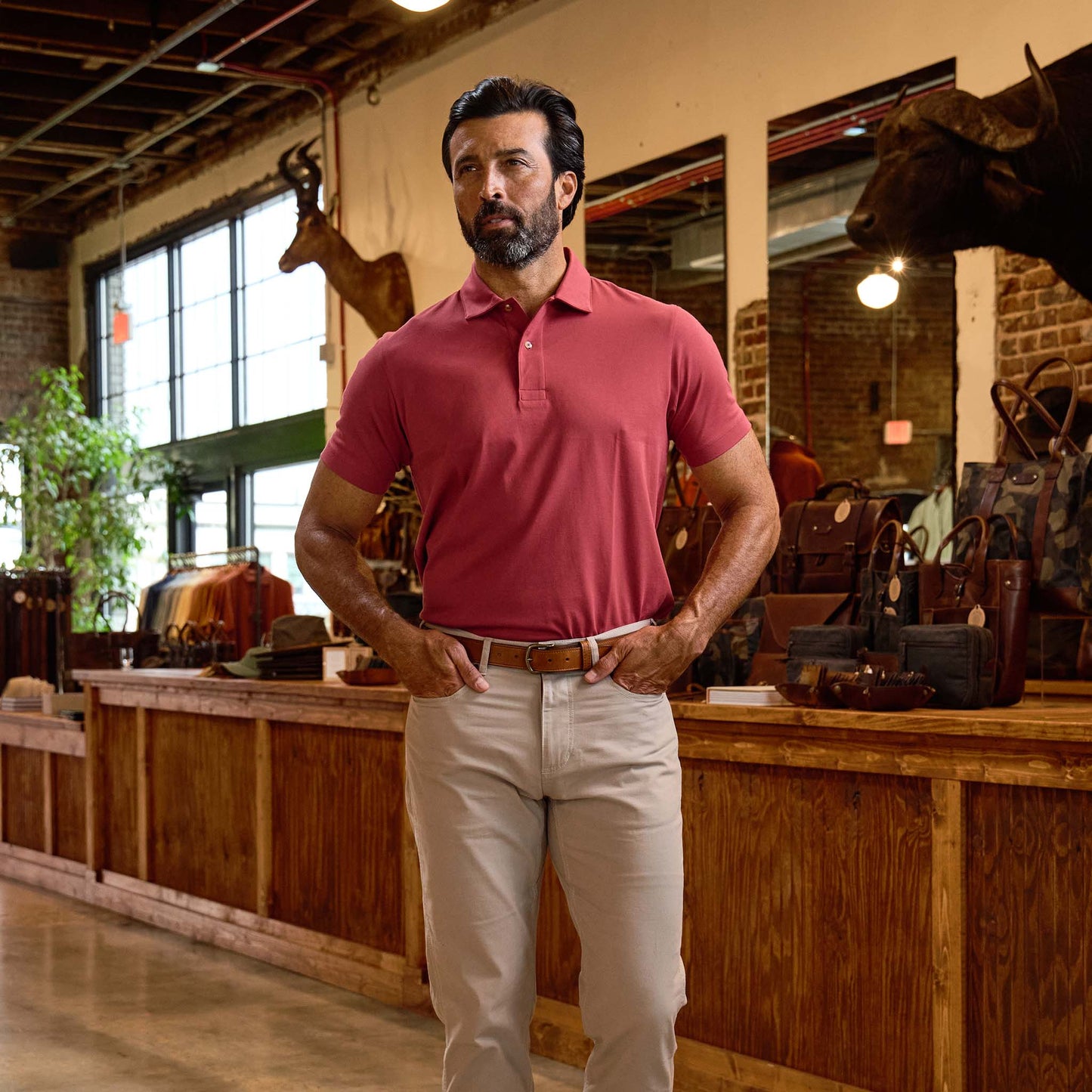 A man in a red polo shirt and beige pants stands in a rustic shop with wooden counters and mounted animal heads.