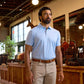 A man in a light blue polo shirt and beige pants stands indoors near wooden shelves and leather bags.