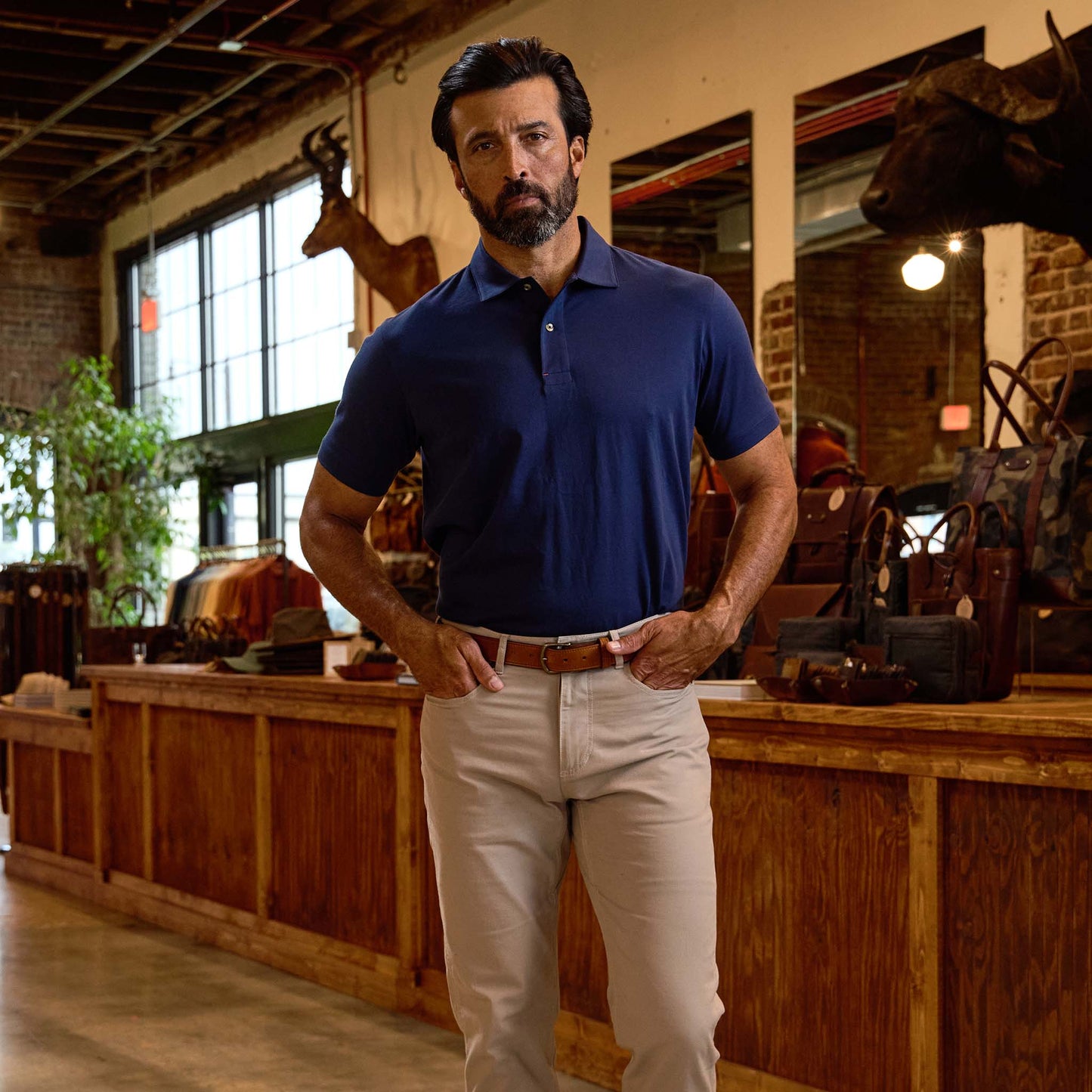 A man in a navy polo shirt and beige pants stands indoors in a store with wood counters and mounted animal heads on the walls.