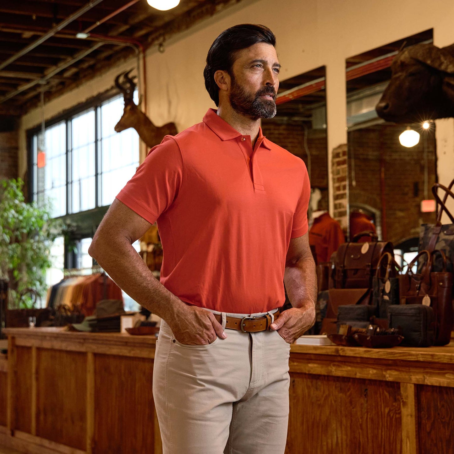Man in orange polo shirt and beige pants stands indoors near wooden counter with bags and mounted animal heads in background.