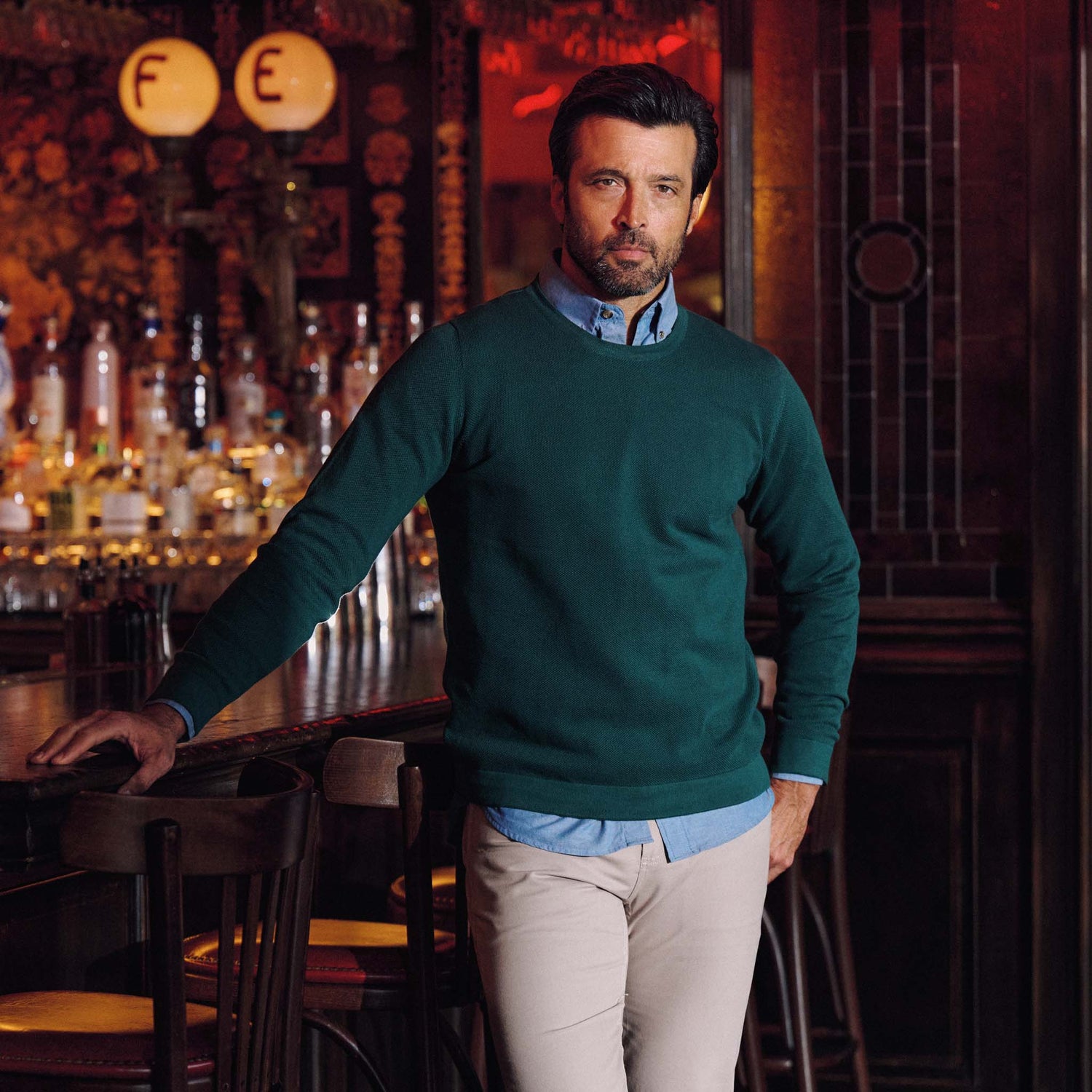 Man in a green Foley Pima Crewneck Sweater by Tom Beckbe and beige pants stands by a warmly lit, inviting bar counter.
