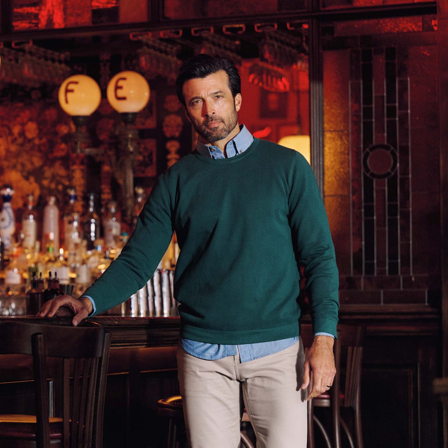 A man in a Tom Beckbe Foley Pima Crewneck Sweater and beige pants stands by a bar with bottles and lights behind him.