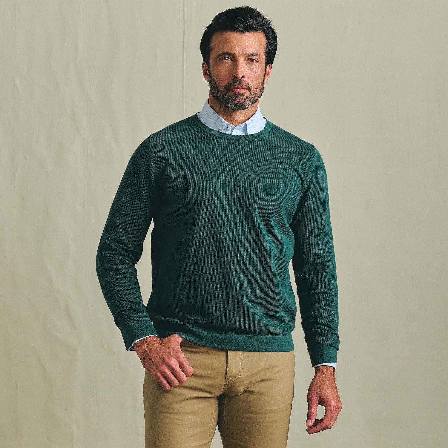 Man wearing the Tom Beckbe Foley Pima Crewneck Sweater over a light blue shirt and tan pants against a plain background.
