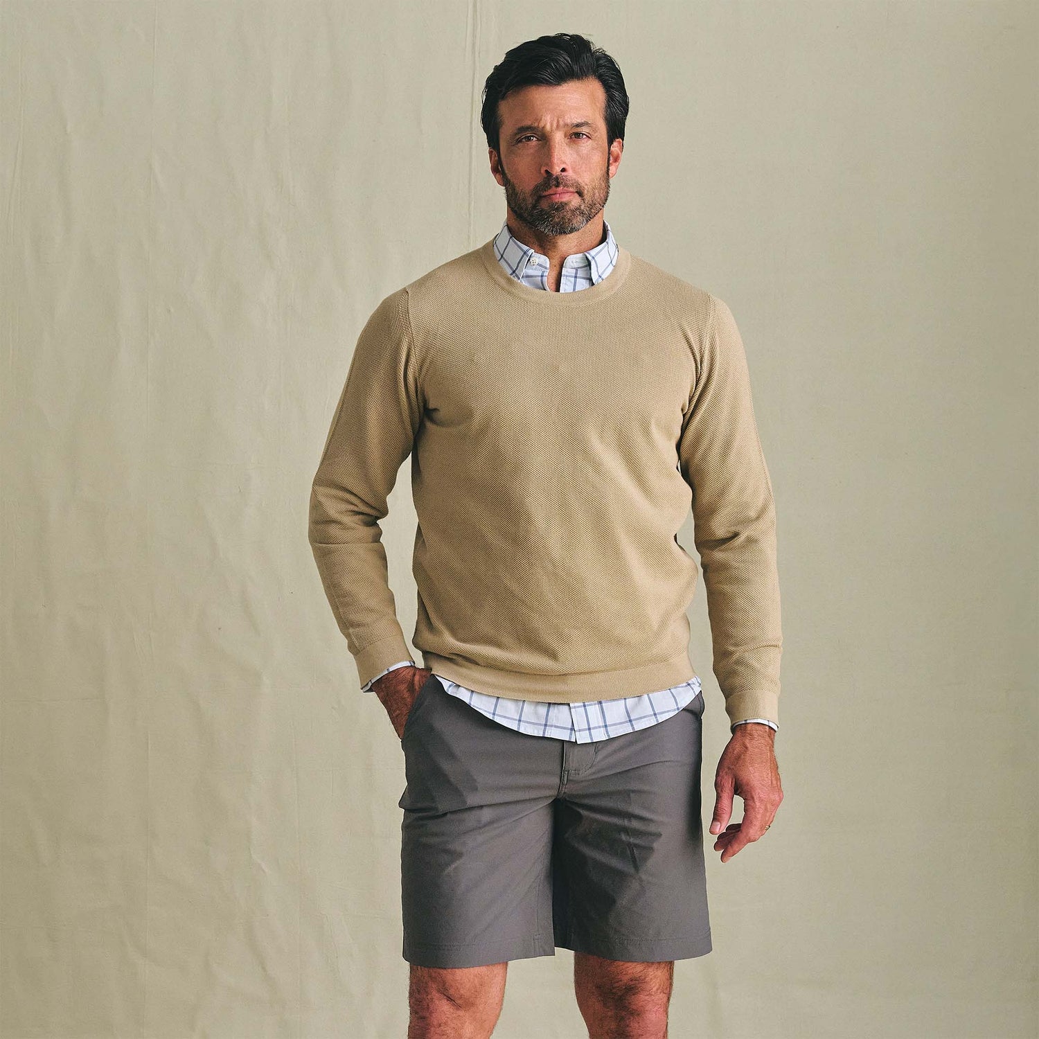A man wears a Tom Beckbe Foley Pima Crewneck Sweater, plaid shirt, and gray shorts against a plain background.