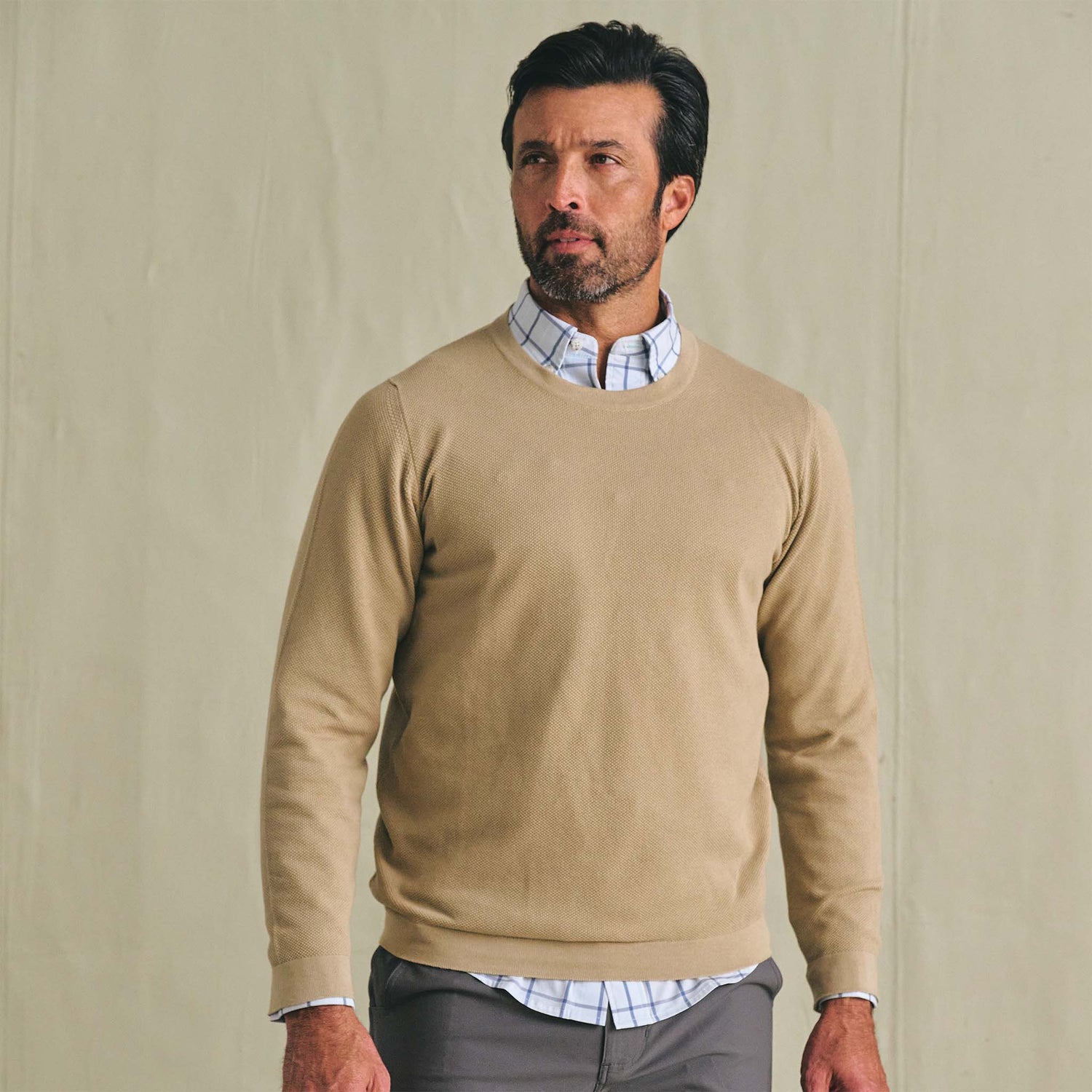 Man wearing the Tom Beckbe Foley Pima Crewneck Sweater over a collared shirt with gray pants, set against a beige background.