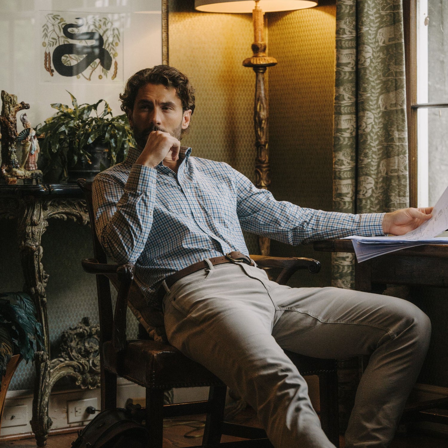 Man in Tom Beckbe’s Powell Performance Shirt sits at a desk, holding papers in a warmly lit, vintage-inspired room.
