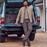 Man in a tan vest and khaki pants leans against the back of an open SUV with travel bags inside on a gravel driveway.