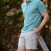 A man in a teal polo and Tom Beckbes Reid Shorts leans against a wooden railing outdoors.