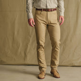 Man wears Tom Beckbe Reid Slim-Straight Leg Cotton Chino Pant, brown belt, and dress shirt against a tan background.