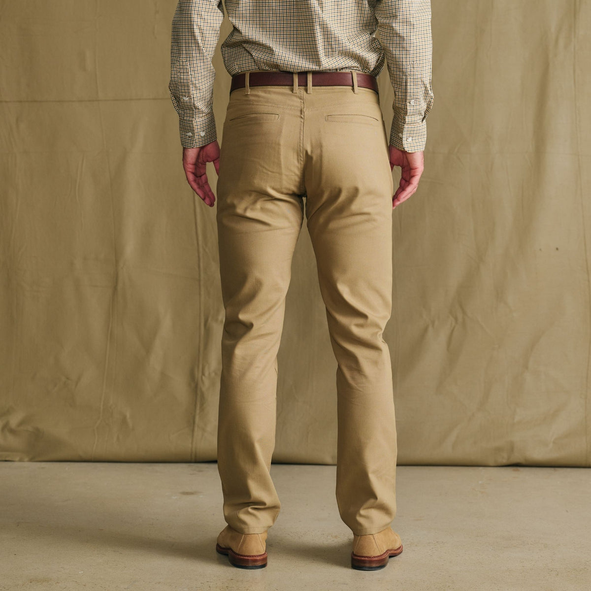 Man wears Tom Beckbe Reid Slim-Straight Leg Cotton Chino Pant, brown belt, and shoes, against beige background.