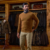 Man wears Tom Beckbe Saunders Crewneck Sweater and khaki pants in a store with jackets and bags behind him.