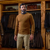 Man in a brown Tom Beckbe Saunders Crewneck Sweater and khaki pants stands before a closet with jackets and bags.