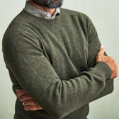 Man wears Tom Beckbe Saunders Crewneck Sweater in olive green over a checkered shirt, arms crossed against plain background.