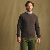 Man wears Tom Beckbe Saunders Crewneck Sweater over a collared shirt with olive pants before a tan backdrop.
