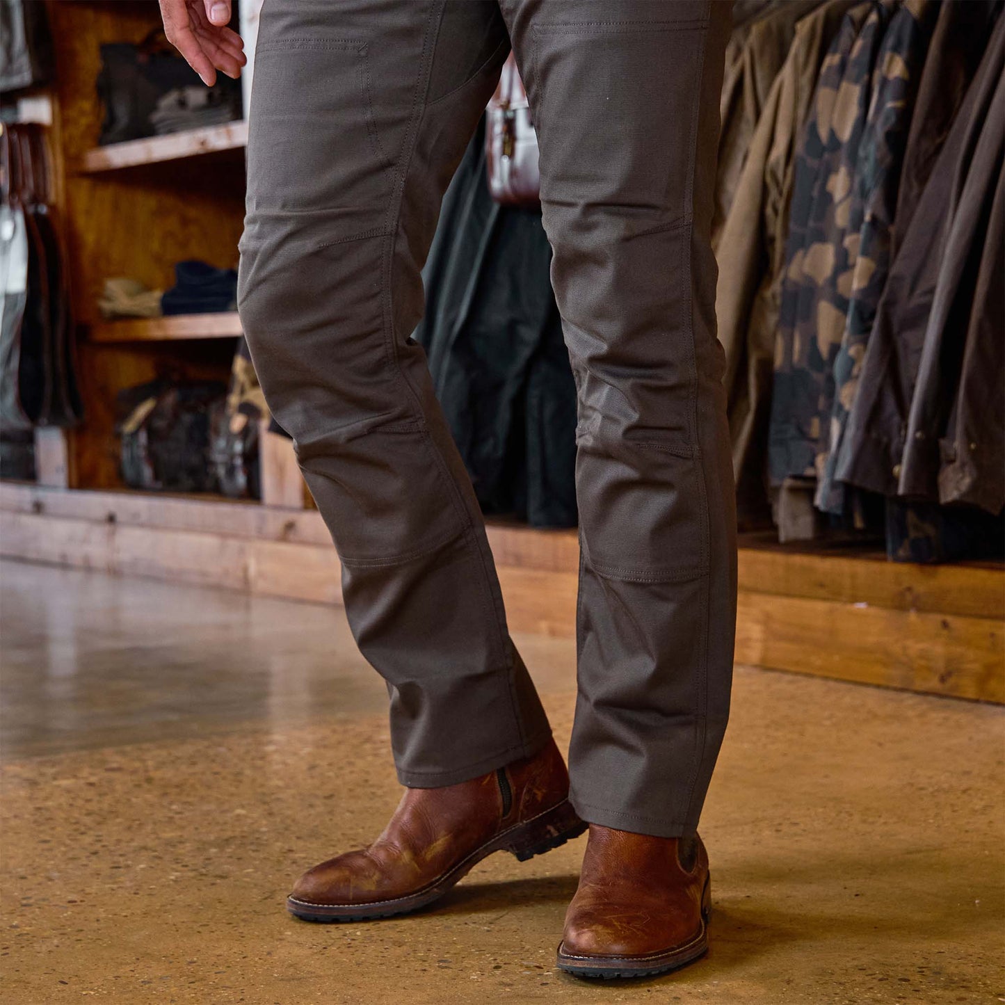 A man wears Tom Beckbe Stafford Double Front Pants and brown leather boots on a polished concrete floor in a store.