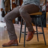 In a store, a person wears Tom Beckbe Stafford Double Front Pants with leather boots, seated on a tall chair.