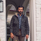 A man in a Tom Beckbe Tensaw Jacket, plaid shirt, and brown pants stands in a white brick doorway.