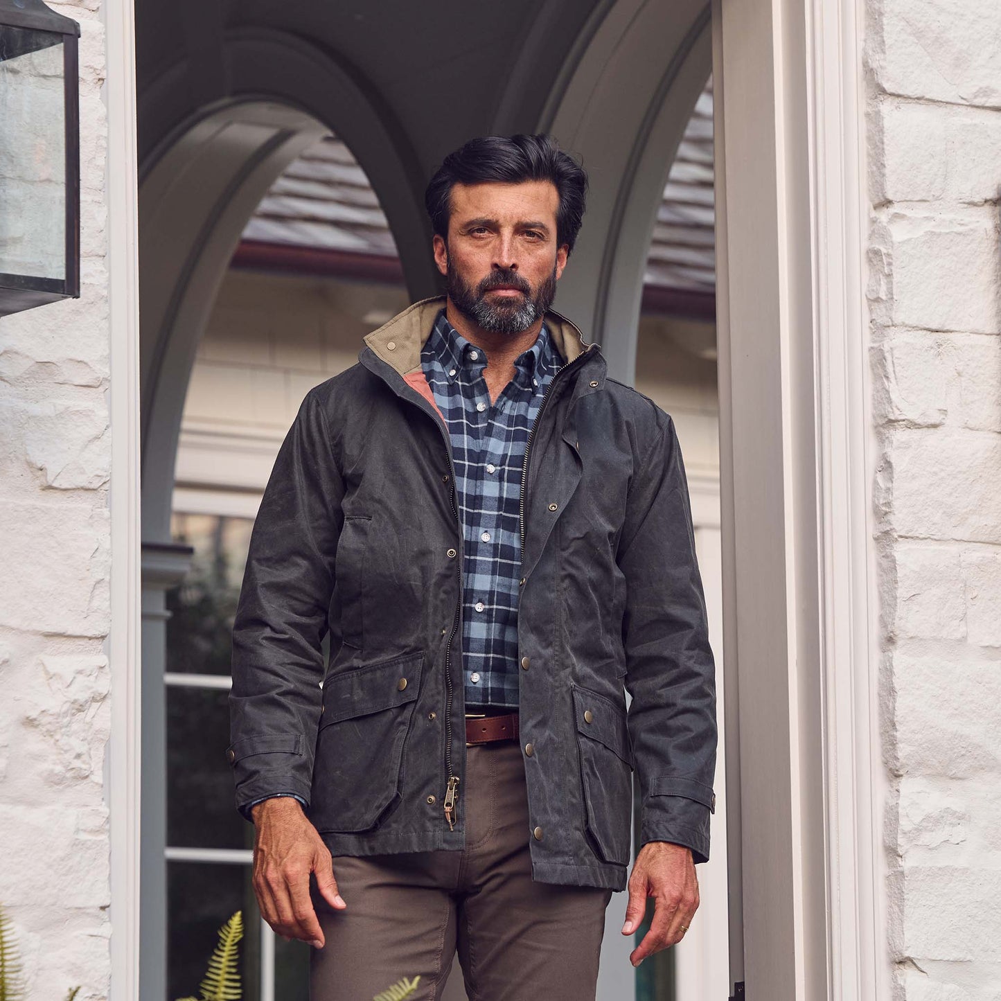 A man in a Tom Beckbe Tensaw Jacket, plaid shirt, and brown pants stands in a white brick doorway.