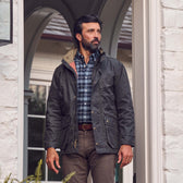 A man in a Tom Beckbe Tensaw Jacket, plaid shirt, and brown pants stands by a light brick wall doorway.