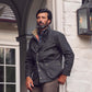 Man with dark hair and beard wears a Tom Beckbe Tensaw Jacket by a white brick wall near a lantern and window.