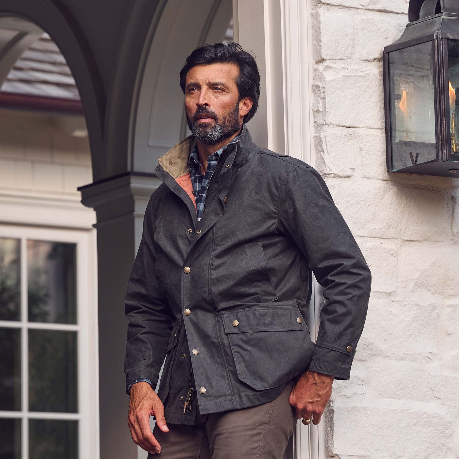 Man with dark hair and beard wears a Tom Beckbe Tensaw Jacket by a white brick wall near a lantern and window.