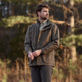 Man wears a brown Tom Beckbe Tensaw Jacket and checkered shirt, standing outdoors by trees, looking to the side.