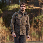 A man in a brown Tom Beckbe Tensaw Jacket and plaid shirt stands outdoors amid trees and foliage.