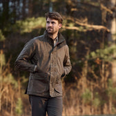 A man in a brown Tom Beckbe Tensaw Jacket and dark pants stands outdoors in the woods, looking to the side.