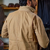 A man in a Tom Beckbe Tensaw Jacket stands indoors by shelves of brown clothing and bags.