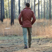 A person in a Fox Red Tensaw Jacket by Tom Beckbe and green pants walks through a forest with tall trees and dry grass.