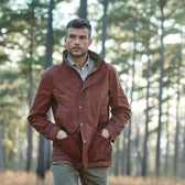 A man in a Tom Beckbe Fox Red Tensaw Jacket stands with hands in pockets against tall forest trees.