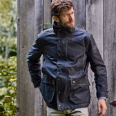Man in a Tom Beckbe Tensaw Jacket with snaps stands by a wooden wall, greenery around.