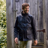 Man in a dark Tom Beckbe jacket smiles, looking aside near a wooden structure outdoors.