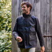 Person outside near a wooden structure, surrounded by greenery, wearing a dark Tom Beckbe Tensaw Jacket.