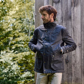 A man in a dark Tom Beckbe Tensaw Jacket stands with hands in pockets, near a wooden wall and lush greenery.