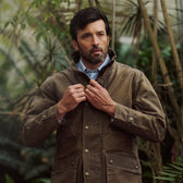 Man outdoors in a Tom Beckbe Tensaw Jacket, fastening the front, surrounded by greenery and rugged style.