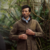 A man outdoors among green plants fastens the buttons on his Tom Beckbe Tensaw Jacket.