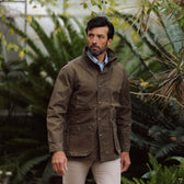 Man wearing a brown Tom Beckbe Tensaw Jacket and light pants stands outdoors with green plants behind him.