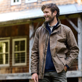 Man wearing a brown Tom Beckbe Tensaw Jacket stands outdoors, smiling with a rustic wooden building in the background.