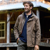 A bearded man stands outside in a Tom Beckbe Tensaw Jacket and dark pants, with a rustic building behind him.
