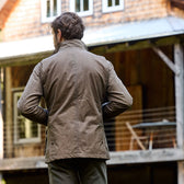 A man in a Tom Beckbe Tensaw Jacket stands outdoors facing a rustic wooden cabin with a porch and railing.