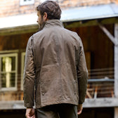 A man in a Tom Beckbe Tensaw Jacket stands with his back to the camera by a rustic wooden building.