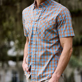 Person wearing a Tom Beckbe Tidewater Shirt (Short Sleeve), blue and brown plaid, outdoors with blurred greenery.