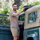 Man in a Tom Beckbe Tidewater Shirt and shorts places a bag on a parked green open-top vehicle in the woods.