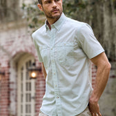 A man in a light blue Tom Beckbe Tidewater short-sleeve shirt stands near a brick building with an arched window.
