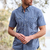 Person wearing a Tom Beckbe Tidewater blue plaid short-sleeve shirt, standing outdoors with trees in the background.