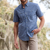 Man outdoors wearing Tom Beckbes Tidewater Shirt (Short Sleeve) in blue plaid with beige pants, trees in the background.