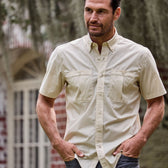 A man outside a brick building with hands in pockets wears a beige, short-sleeve Tidewater Shirt by Tom Beckbe with UPF 30+.