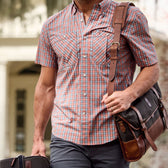 A person in a Tidewater Shirt by Tom Beckbe carries a black-brown messenger bag and a small suitcase while walking outside.